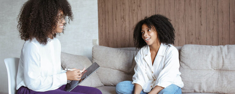 A black woman wearing a a white button up shirt sits cross legged on a beige couch across from another black woman in a white blouse who is holding a tablet.