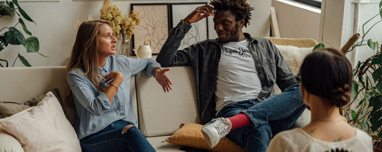 An interracial couples speaks to one another as they sit on a couch across from a therapist.