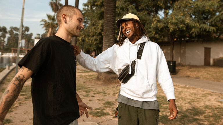 A man with locs wearing a hat, white hoodie, and cross-body bag laughs and places his hand on the shoulder of a man who has his head shaved and is wearing a black t-shirt.