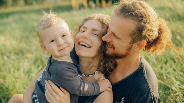 A fair-skinned woman sitting in a grassy field holds her baby out in front of her while her husband embraces her from behind.