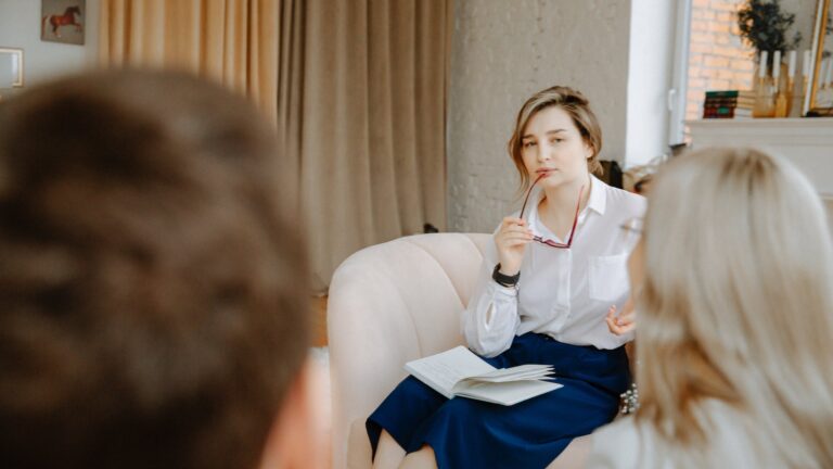 Therapist is framed between the back of the heads of two clients; she's looking at them while clutching her glasses in her hand and resting them near her mouth.