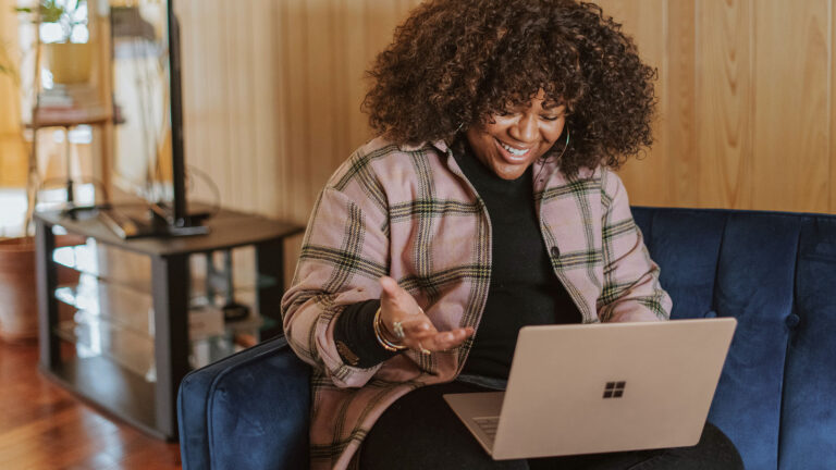 A woman in a plaid jacket sits on a blue couch while attending a zoom call on the laptop in her lap.