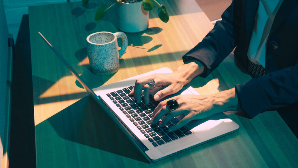 A woman, wearing a blazer, large black ring, and face unseen, types at a laptop. computer