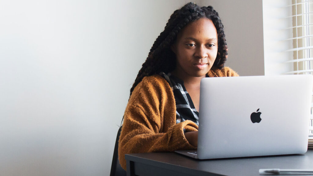 A Black woman wearing a yellow cardigan sits at a table working on a laptop.