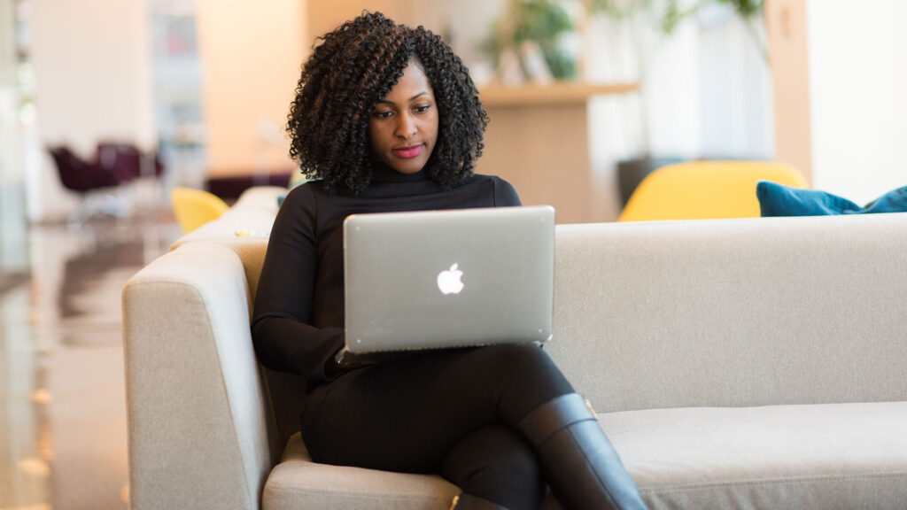 A Black woman sits on a tan couch holding a Macbook in her lap.