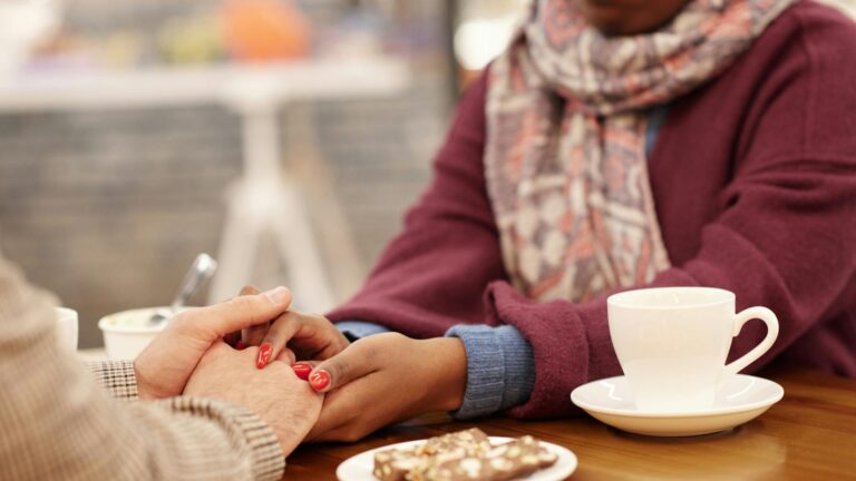 Black woman with red painted nails holding the hands of a white man over pastries and coffee.