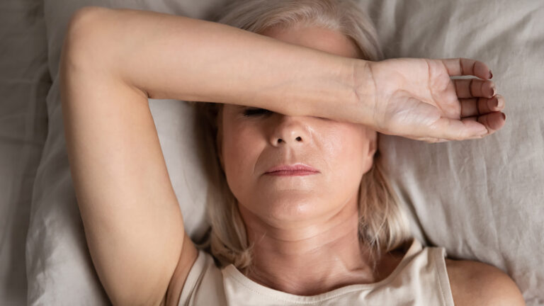 White woman with beige sleeveless shirt rests her arm across her eyes while she lies on a bed looking worn out