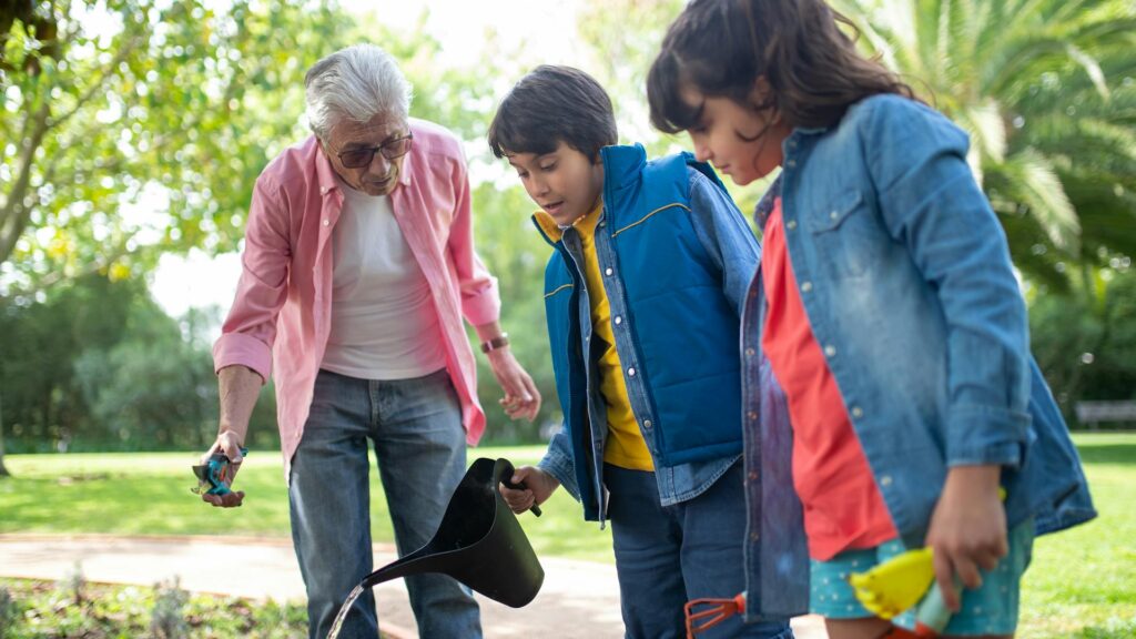 A Latino boy with a blue vest on waters a garden with a black watering can as he stands between his grandpa in a pink button up shirt holding a garden tool and his sister in a denim shirt holding other garden tools.