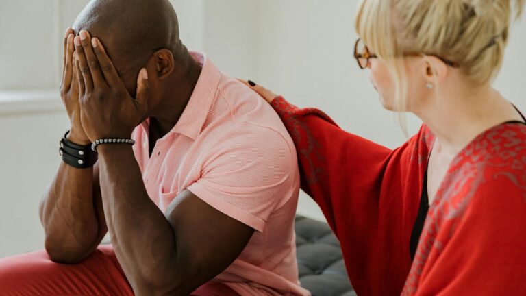 A white woman in a red shirt places a comforting arm on the shoulder of a black man in an orange shirt. The man is covering his face with his hands in a sign of distress.