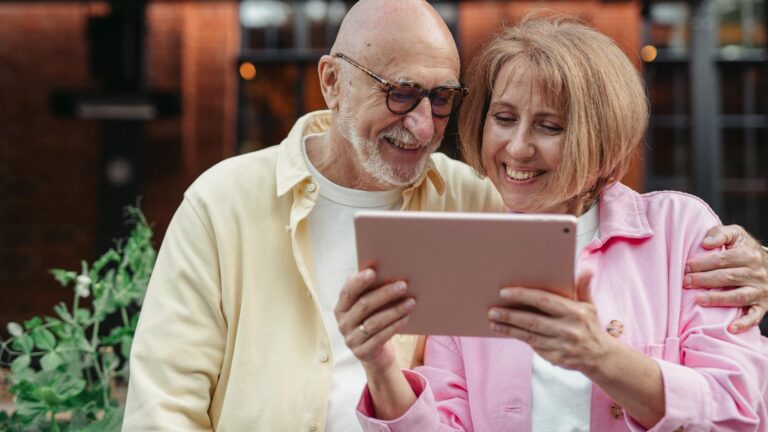 A while elderly couple looks at a pink iPad. The man is on the left, wearing glasses and a yellow button-up. THe woman is holding the iPad with two hands and wearing a pink button-up.