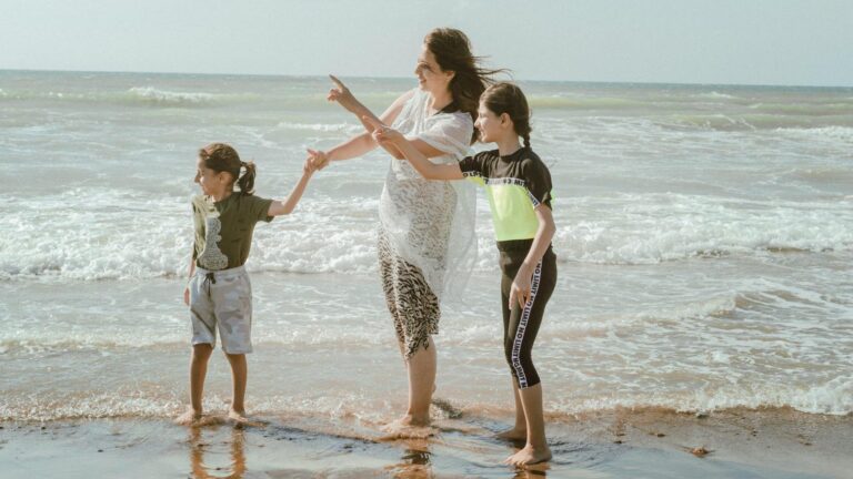 A woman in a flowy white dress stands on the shoreline of a beach with her two children.