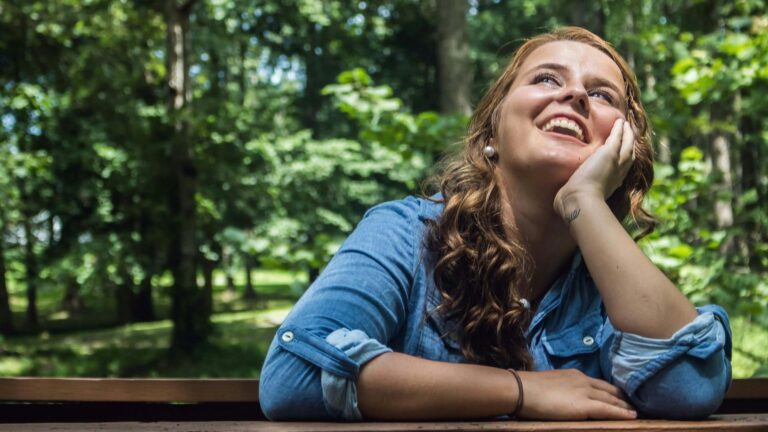 White woman in denim shirt with sleeves rolled up rests her chin in her hand and looks up at the sky.