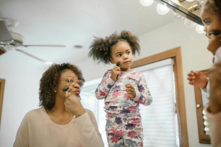 An older black woman and a black female toddler look into a mirror while using a stone roller on their faces for skincare.