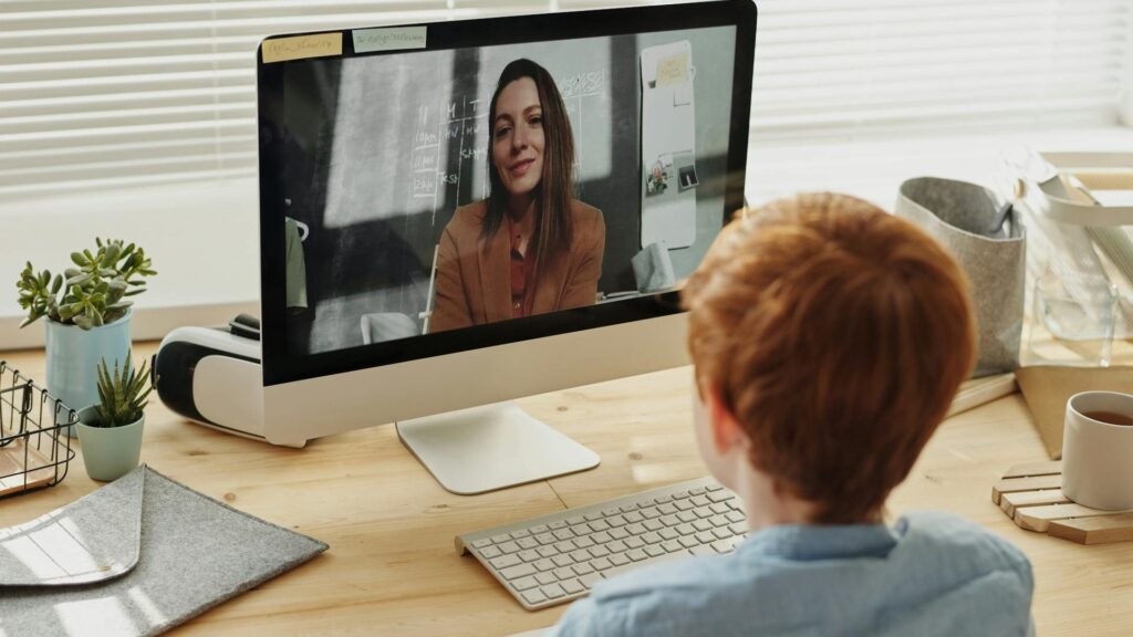 A therapist is seen on a computer screen during a telehealth therapy appointment speaks to a young boy with red hair.