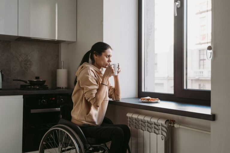 A brunette, wheelchair bound woman wearing a tan hoodie sips coffee from a white mug as she looks out the kitchen window in her city apartment.