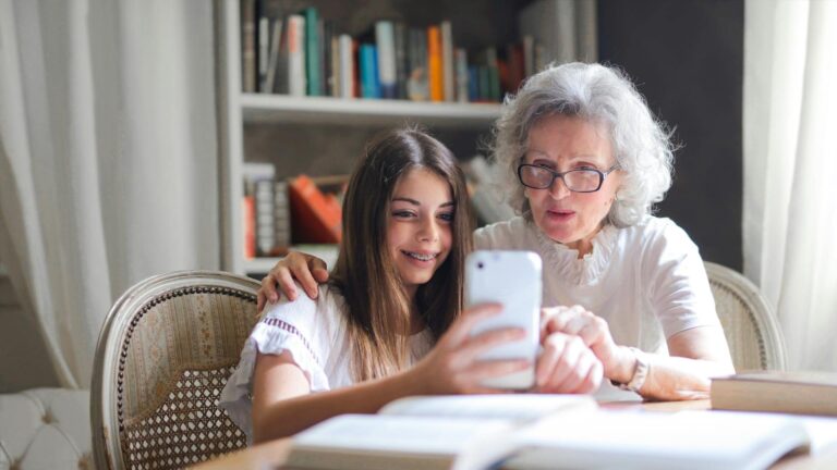 A young girl with long brown hair and braces sits at a dining table covered with books next to an elderly woman with grey hair. The girl is holding up a cell phone, showing the older woman something on the phone.