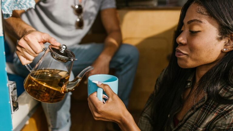 A Black woman in a plaid shirt holds a blue mug as a man pours tea into her mug.