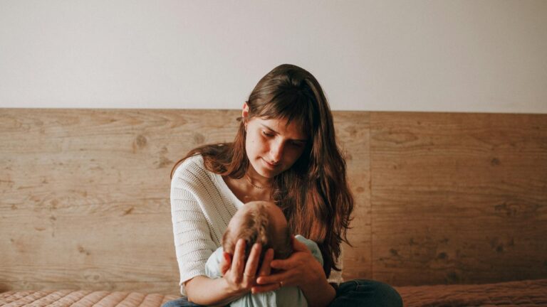 A white woman with long brown hair wearing a white mid-sleeve top sits on a bed with her legs crossed while she holds a baby’s head in her hands and looks down at the baby’s face.