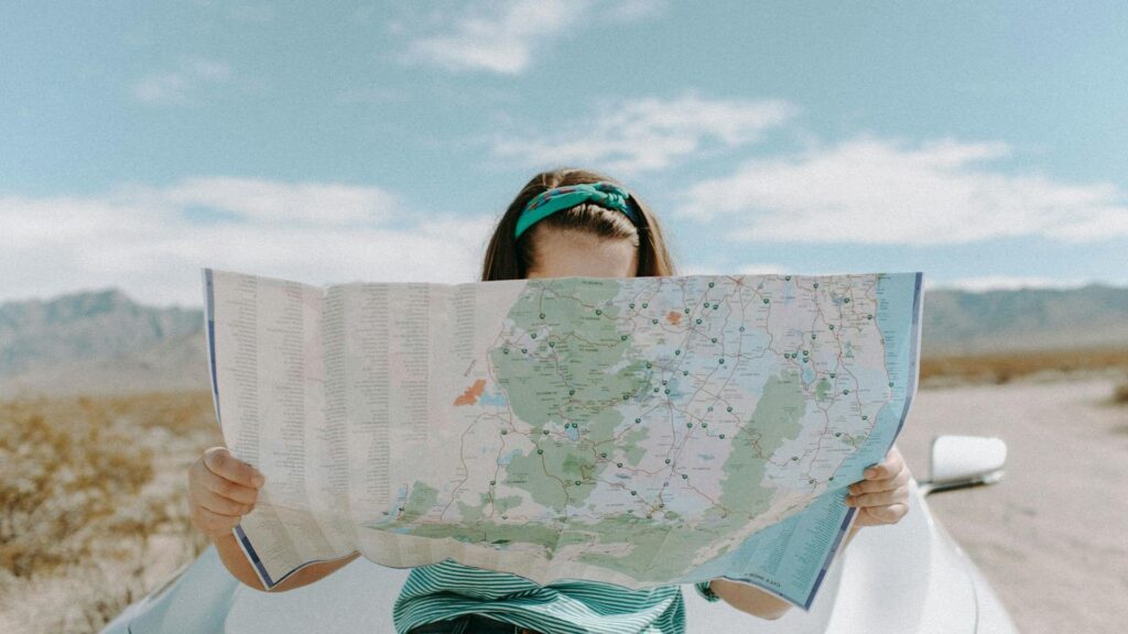 A white woman holds a large map in front of her face as she stands in front of a silver vehicle.
