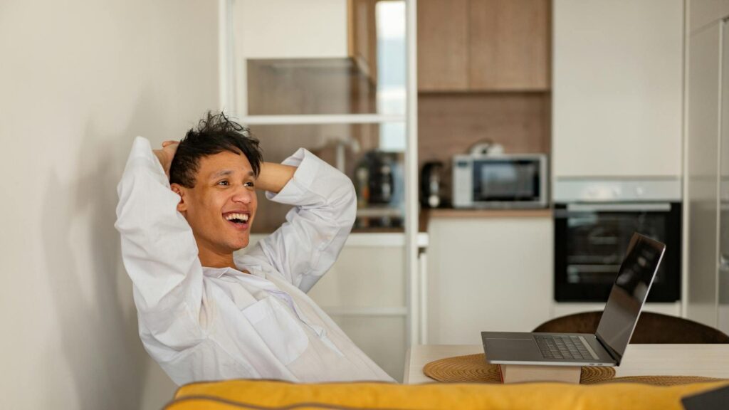 A smiling man with black hair sits in an apartment in front of a laptop with his hands on his head, looking off in amused disbelief.