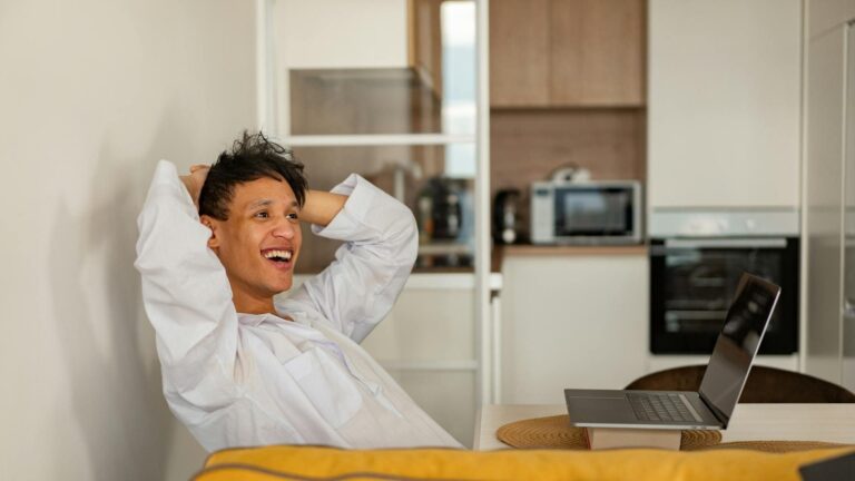 A smiling man with black hair sits in an apartment in front of a laptop with his hands on his head, looking off in amused disbelief.