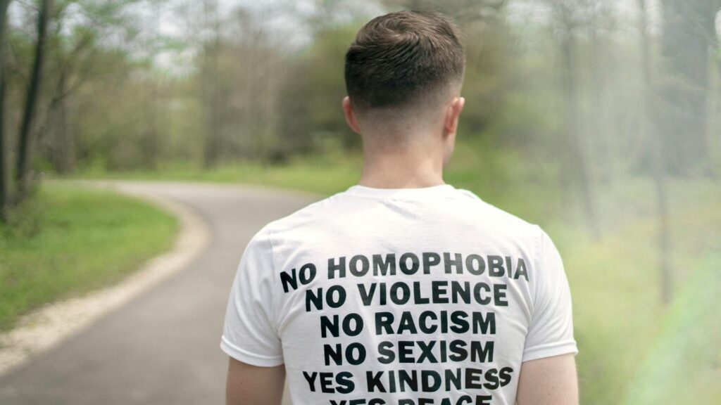 A white man wearing a white t-shirt with the words,”no homophobia no violence no racism no sexism yes kindness yes peace yes equality yes love,” walks away from the camera down a paved path through a wooded area.