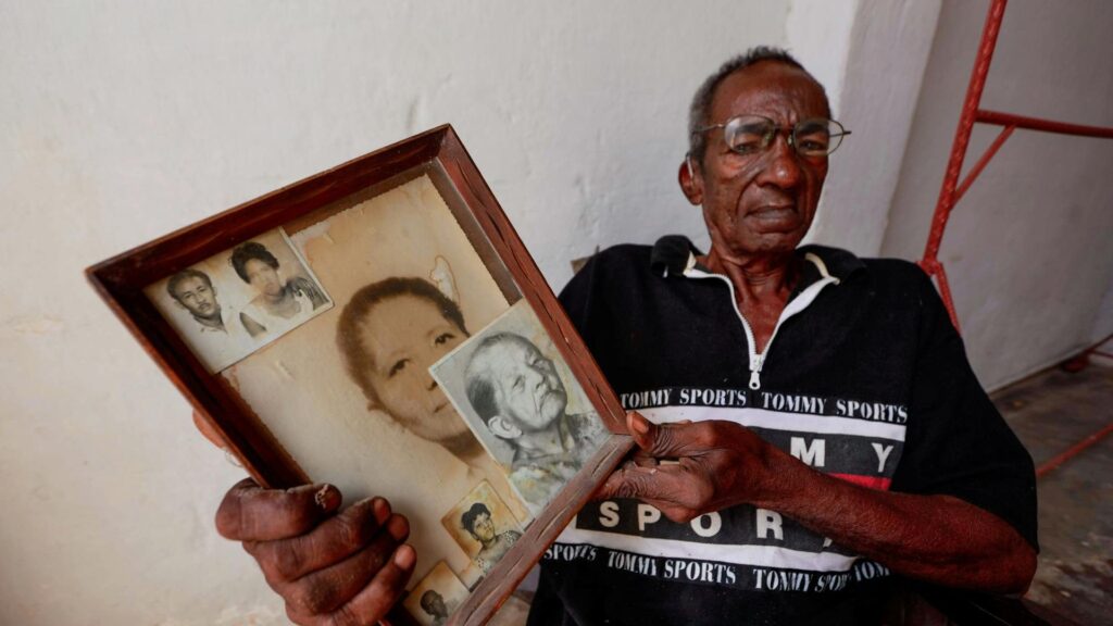 A Black man in glasses sits in a chair while holding up several vintage portraits of his family members.