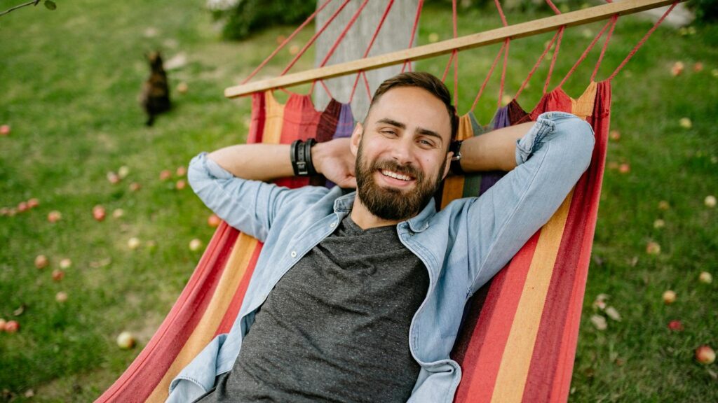A bearded man rests his arms behind his head as he reclines in a colorful hammock.
