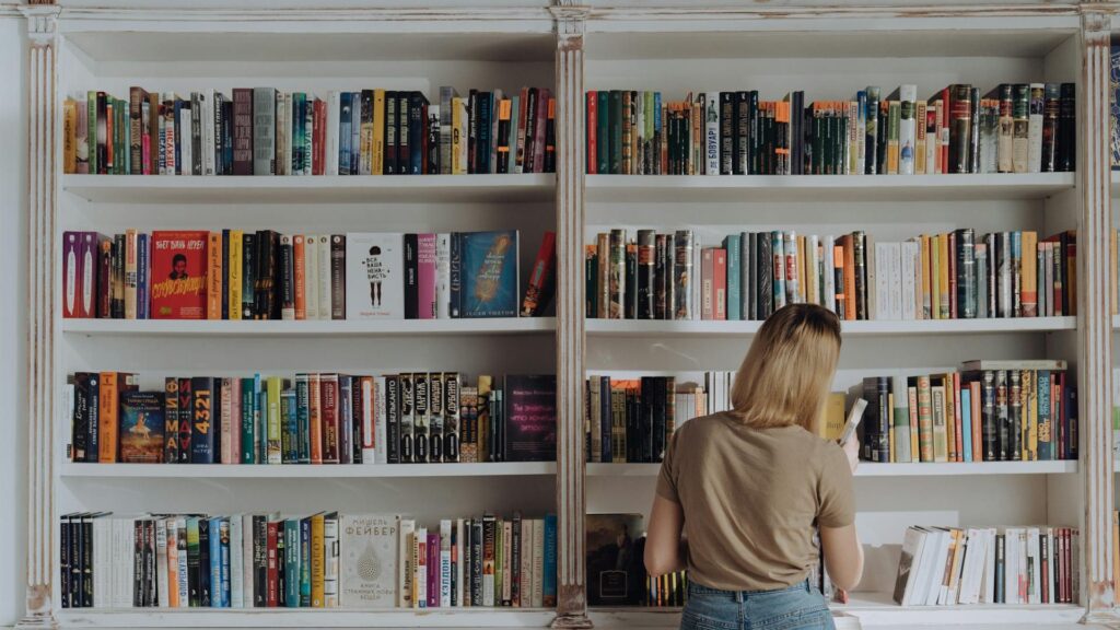 A woman with shoulder-length blonde hair, wearing a beige t-shirt and denim skirt, is standing in front of a large white bookshelf filled with colorful books. She is facing away from the camera, browsing the selection, and holding a book in her right hand.