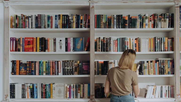 A woman with shoulder-length blonde hair, wearing a beige t-shirt and denim skirt, is standing in front of a large white bookshelf filled with colorful books. She is facing away from the camera, browsing the selection, and holding a book in her right hand.