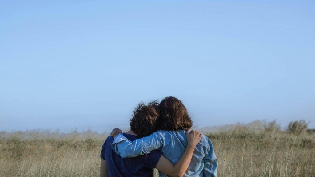A woman is shown in two contrasting scenes within the same home. On the left, she stands in a warmly lit kitchen, casually holding a banana, dressed in a hoodie and loose pants. On the right, she appears in a dimly lit blue-toned room, standing still with a solemn expression, creating a stark contrast in mood and atmosphere.