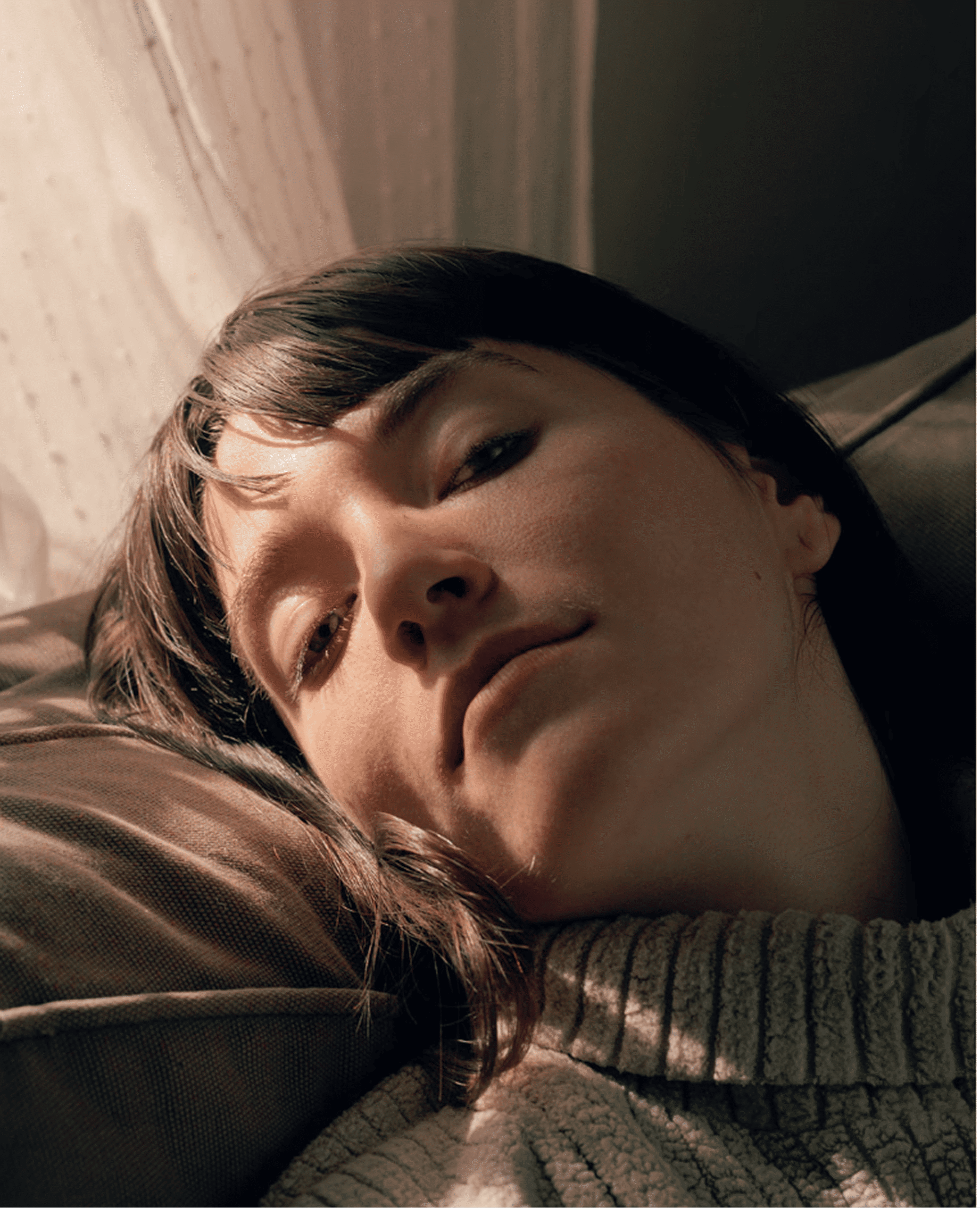 Young woman with dark hair reclines on a couch in soft light. She wears a textured sweater, evoking a calm, intimate atmosphere.