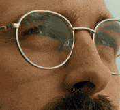 Close-up image of a person wearing round glasses, looking thoughtfully into the distance. The sunlight reflects softly on the lenses, creating a contemplative mood.