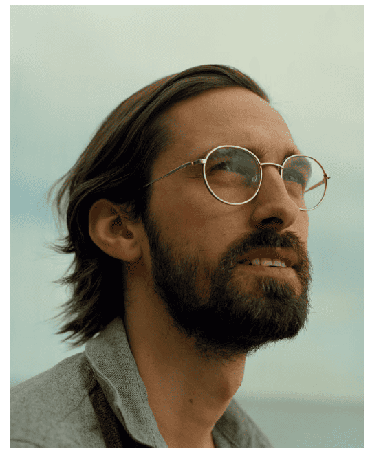 A bearded man with glasses looks thoughtfully upwards against a pale blue sky. He has long hair, creating a calm and contemplative mood.