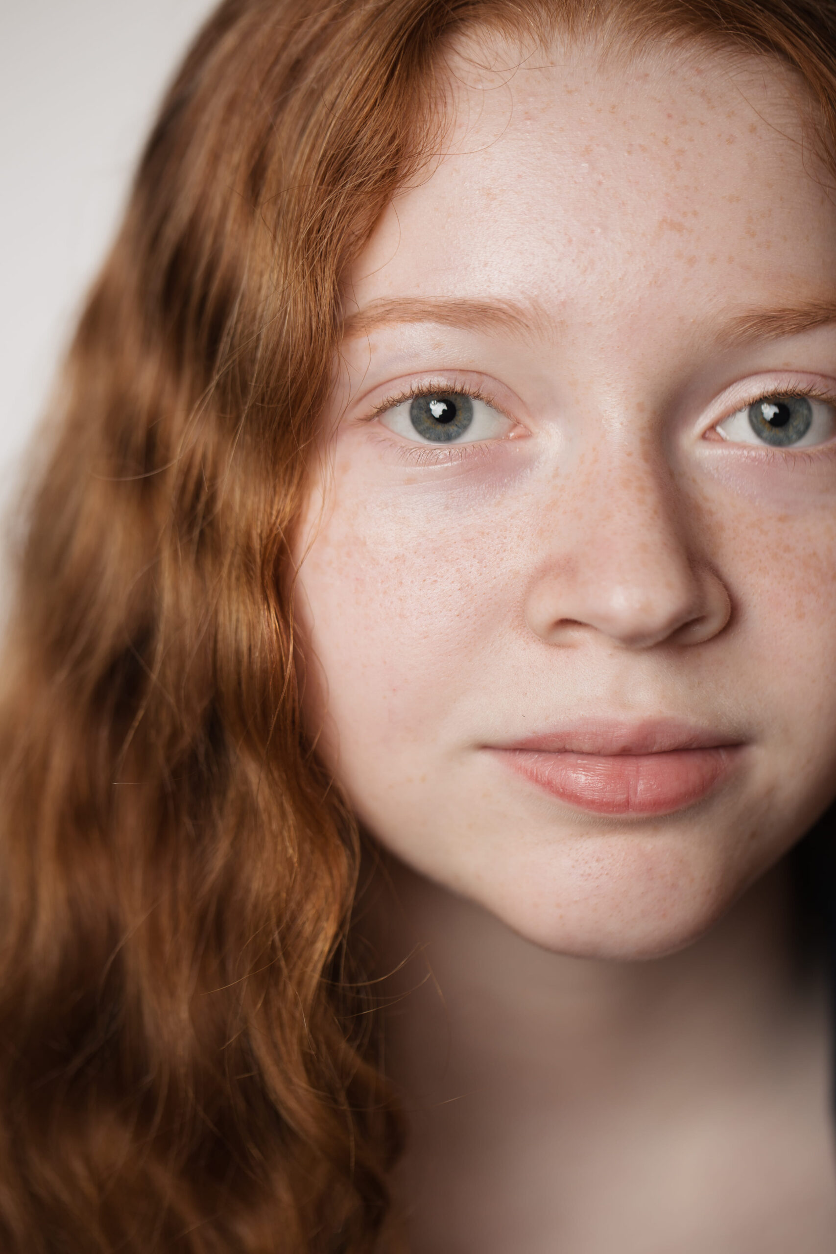 Close-up of a young person with long, wavy red hair, light skin with freckles, and an expression of calmness. The background is softly blurred.