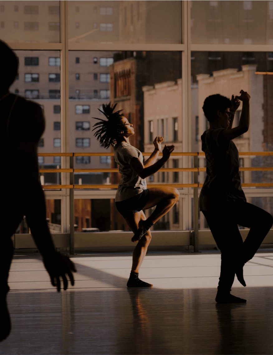 Dancers practice in a sunlit studio, silhouetted by large windows with an urban backdrop. Energetic movements create a dynamic and focused atmosphere.