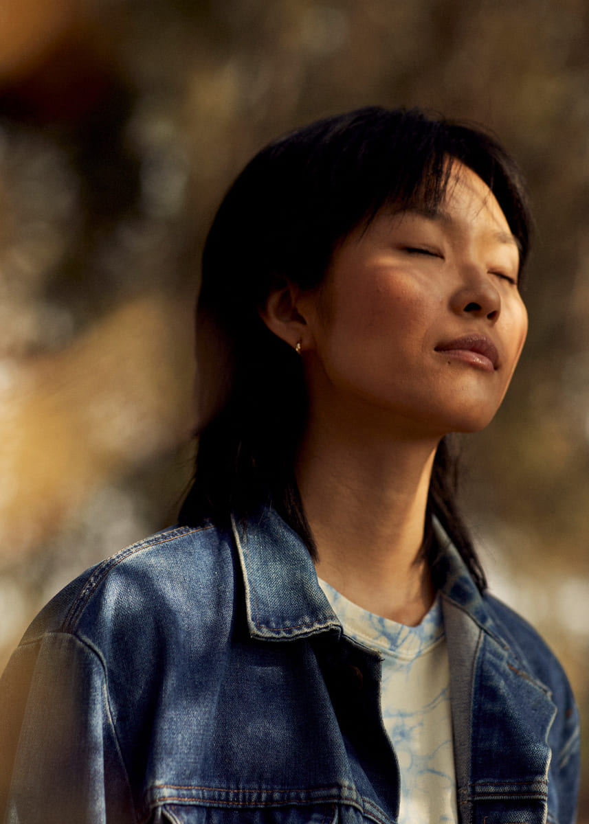 A person with closed eyes stands peacefully in a sunlit forest, wearing a denim jacket. The scene conveys serenity and introspection.
