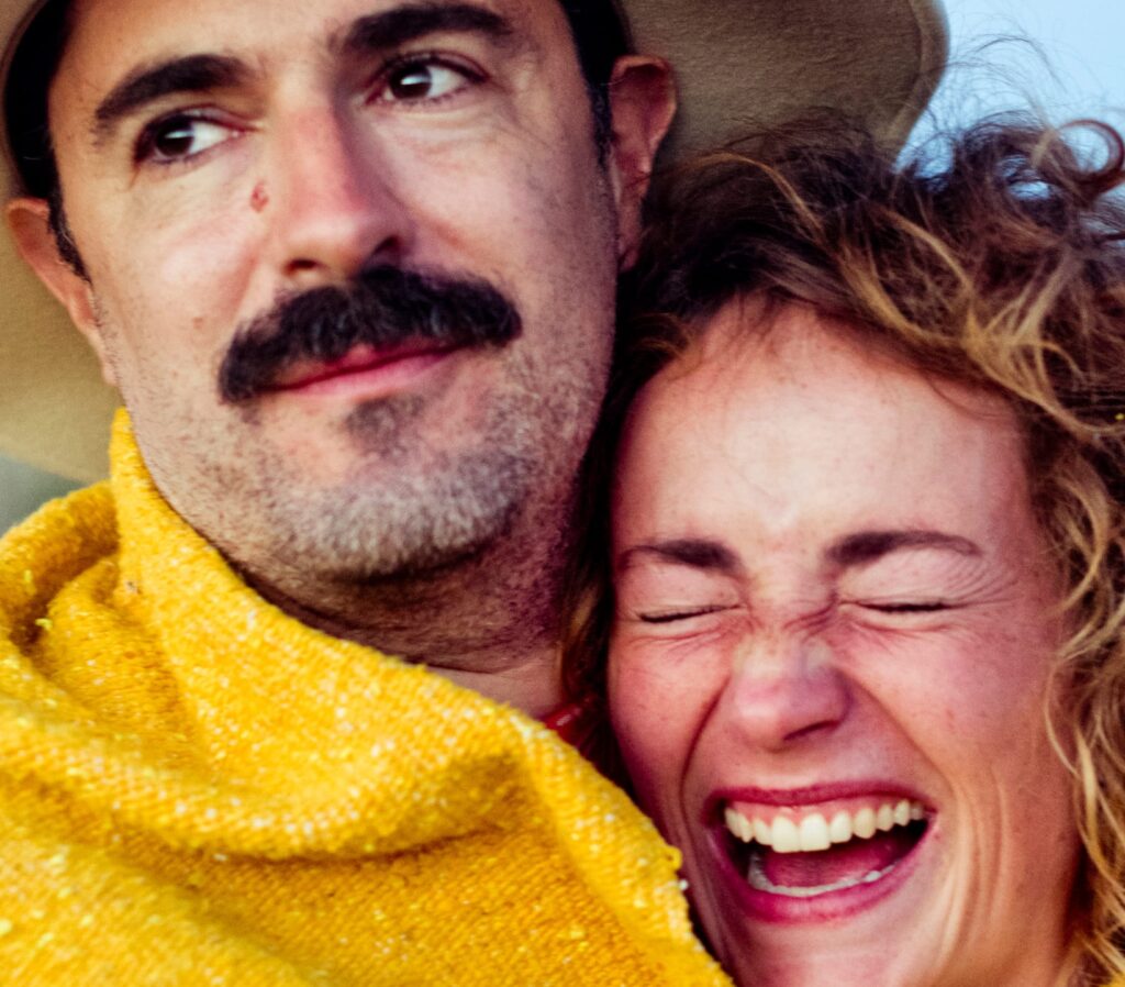 A man with a mustache wearing a hat and a yellow blanket smiles softly while a woman beside him laughs joyfully, eyes closed, conveying happiness.