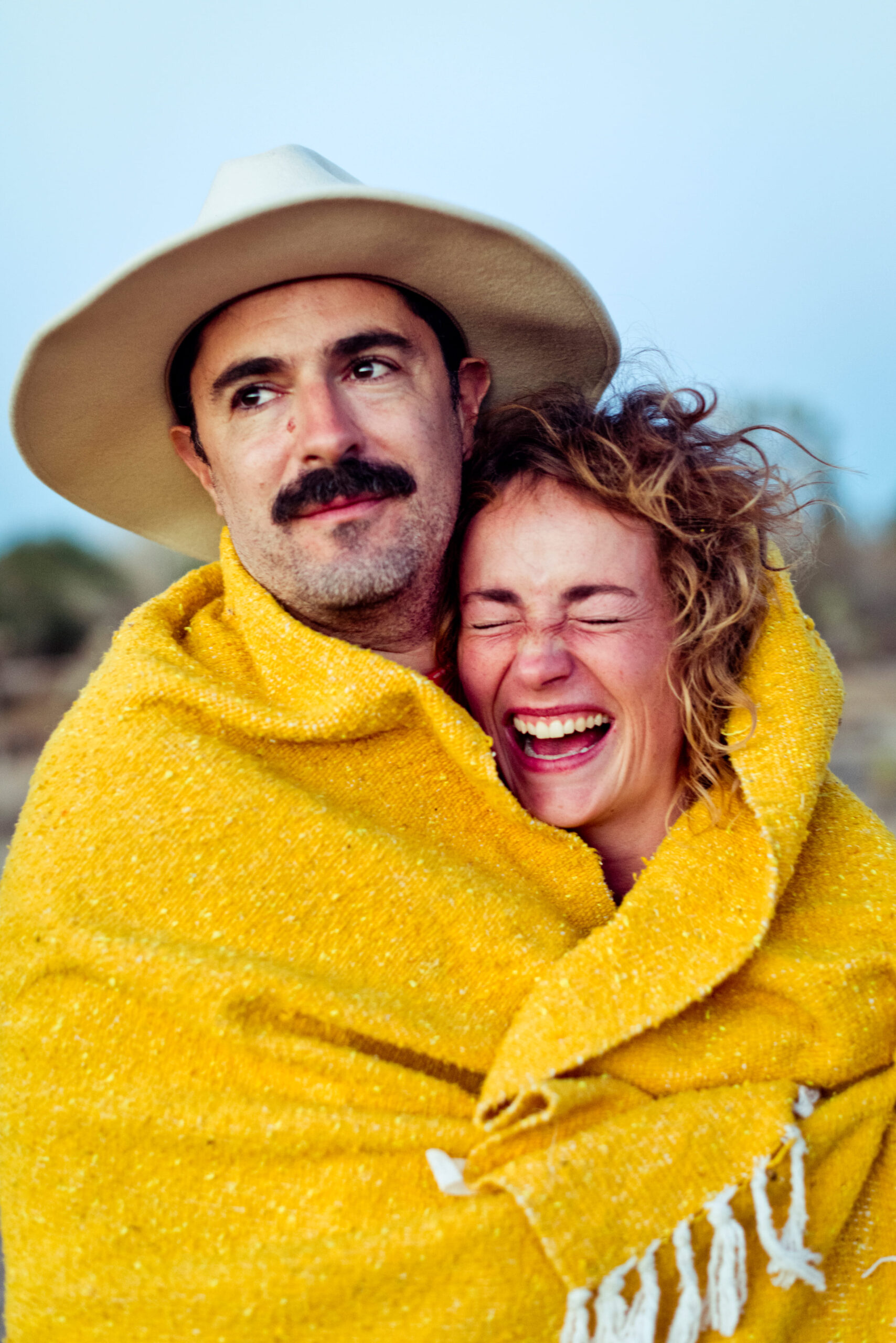 A man in a cowboy hat and a woman with closed eyes and a big laugh are wrapped in a yellow blanket, exuding warmth and joy in an outdoor setting.