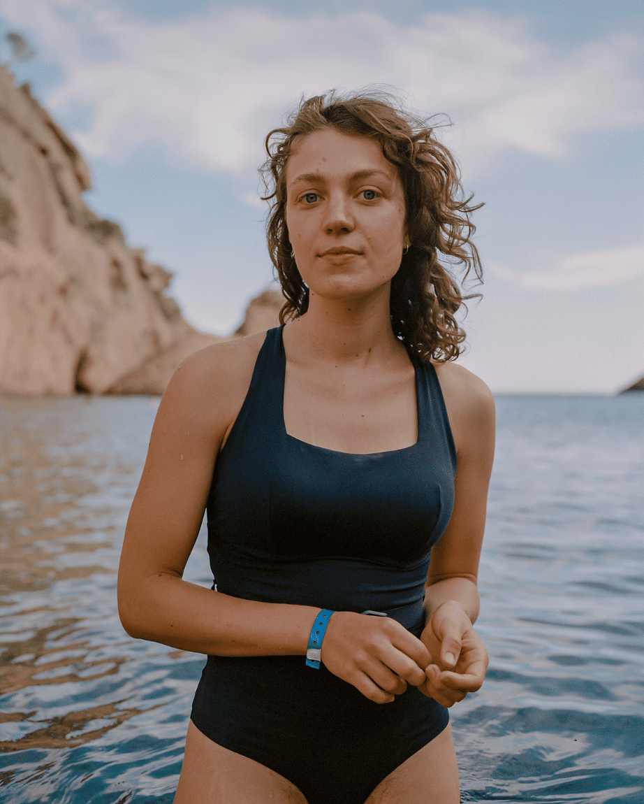 Young woman in a navy swimsuit stands waist-deep in calm ocean water, with rocky cliffs in the background. She gazes confidently at the camera.