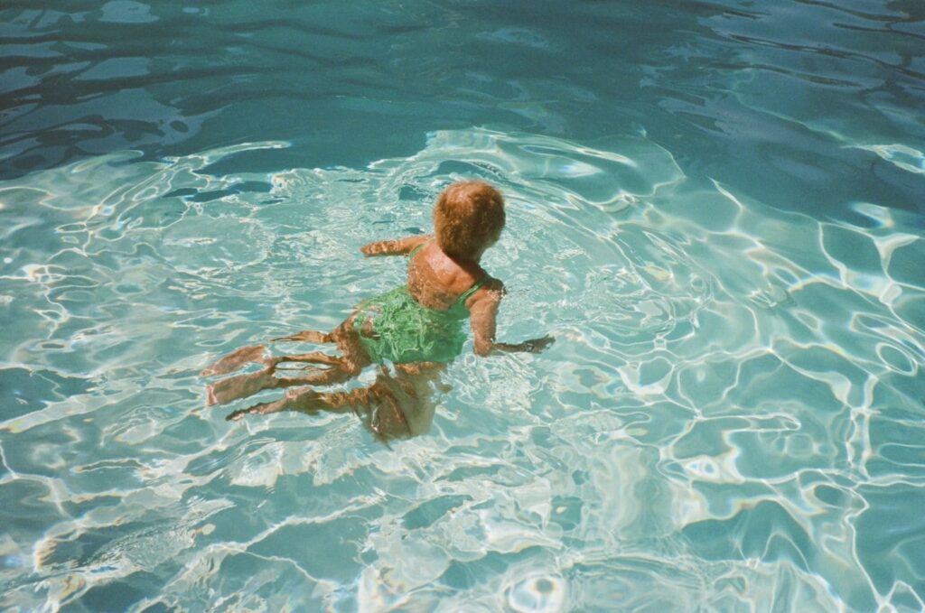 A child in a green swimsuit floats in clear blue water. Sunlight creates shimmering patterns on the surface, conveying a sense of joy and relaxation.