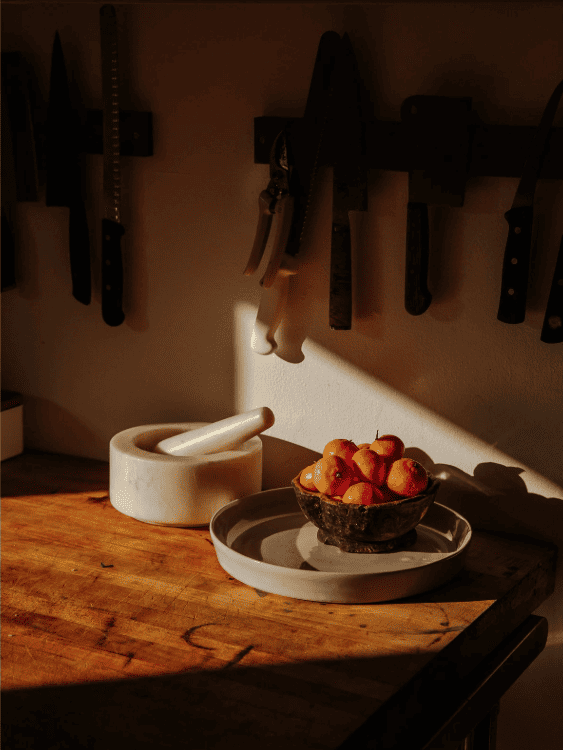 A sunlit kitchen scene with knives on a wall rack, a marble mortar and pestle, and a bowl of tomatoes on a plate, creating a warm, rustic atmosphere.