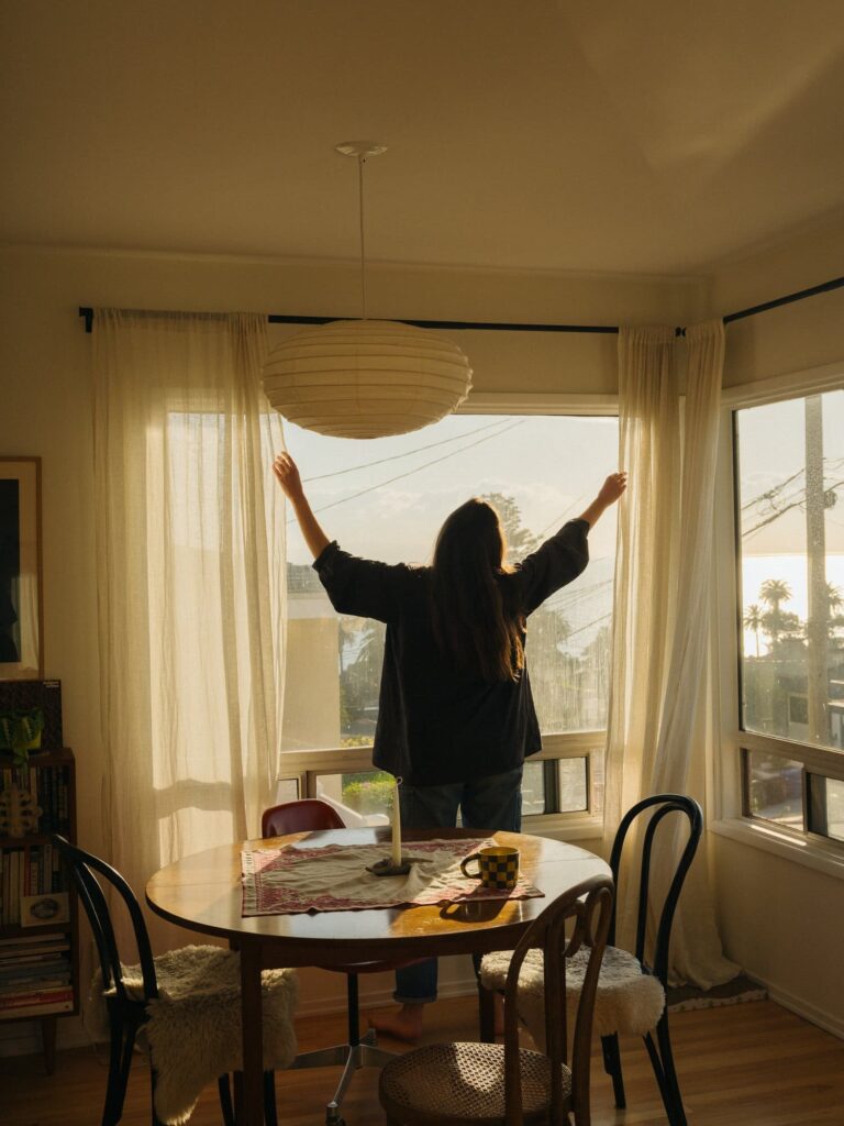 A person stands barefoot, opening curtains in a sunlit room. A dining table with a mug and centerpiece is in the foreground, evoking a cozy morning vibe.