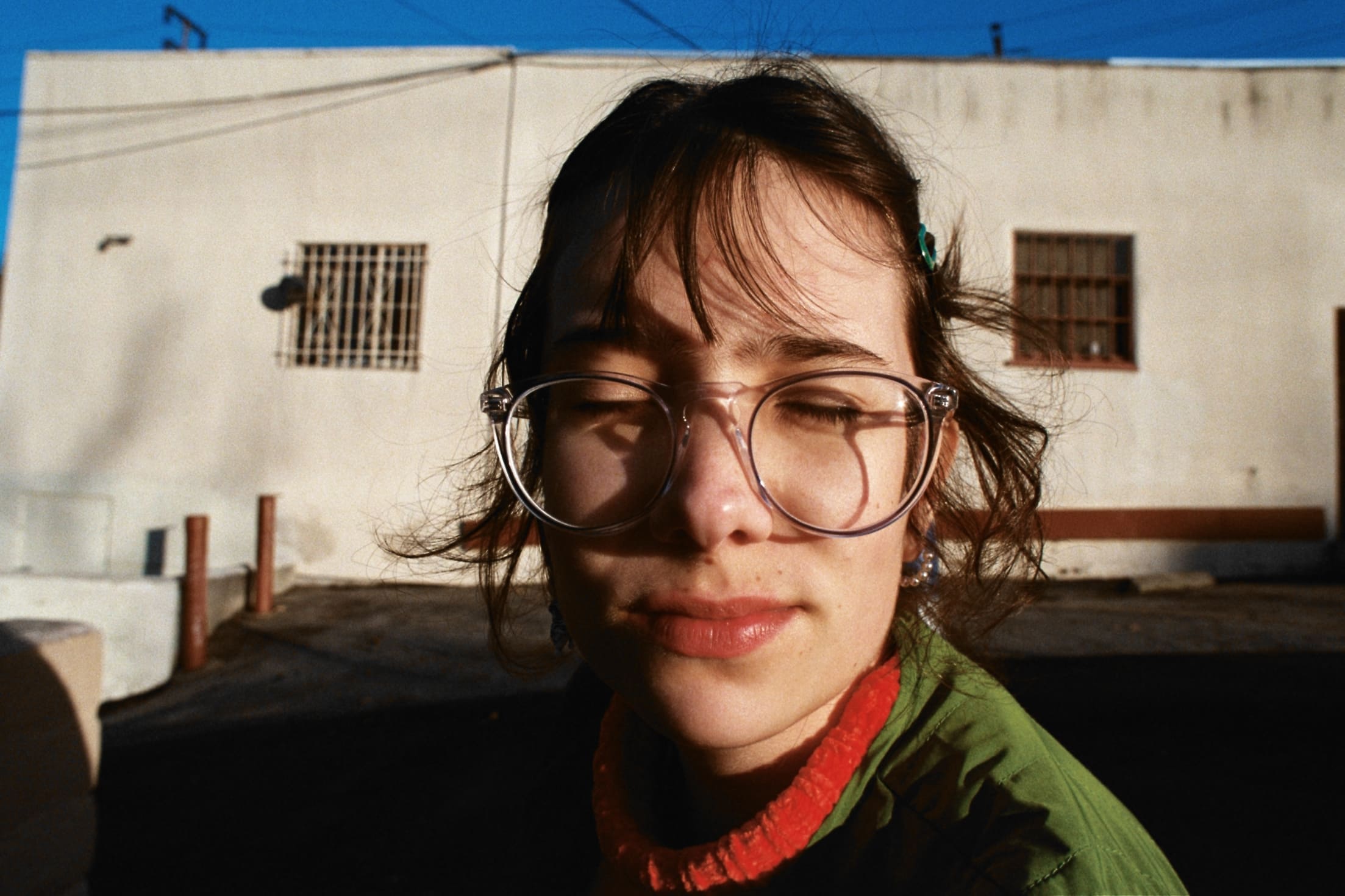 Young person with large glasses, eyes closed, in sunlight. Wearing green jacket, red collar. Plain building, blue sky in background. Relaxed mood.