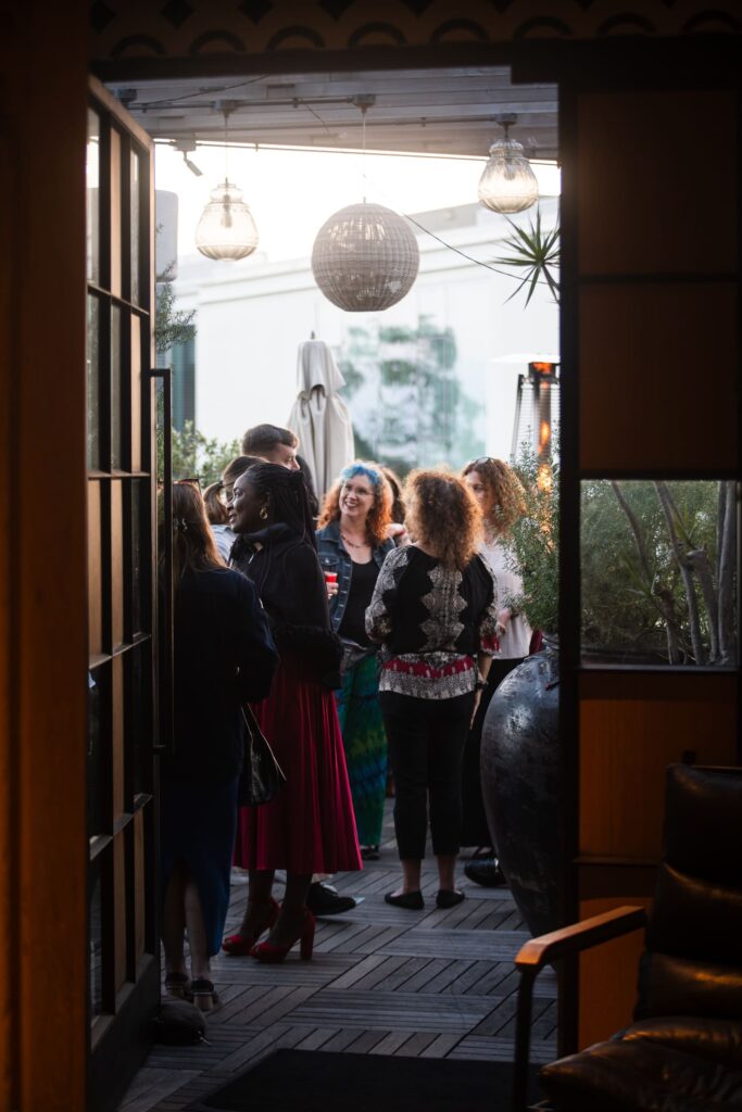 A group of people socializing on a stylish patio with warm evening lighting. Hanging lights and a large pot add a cozy, inviting ambiance.