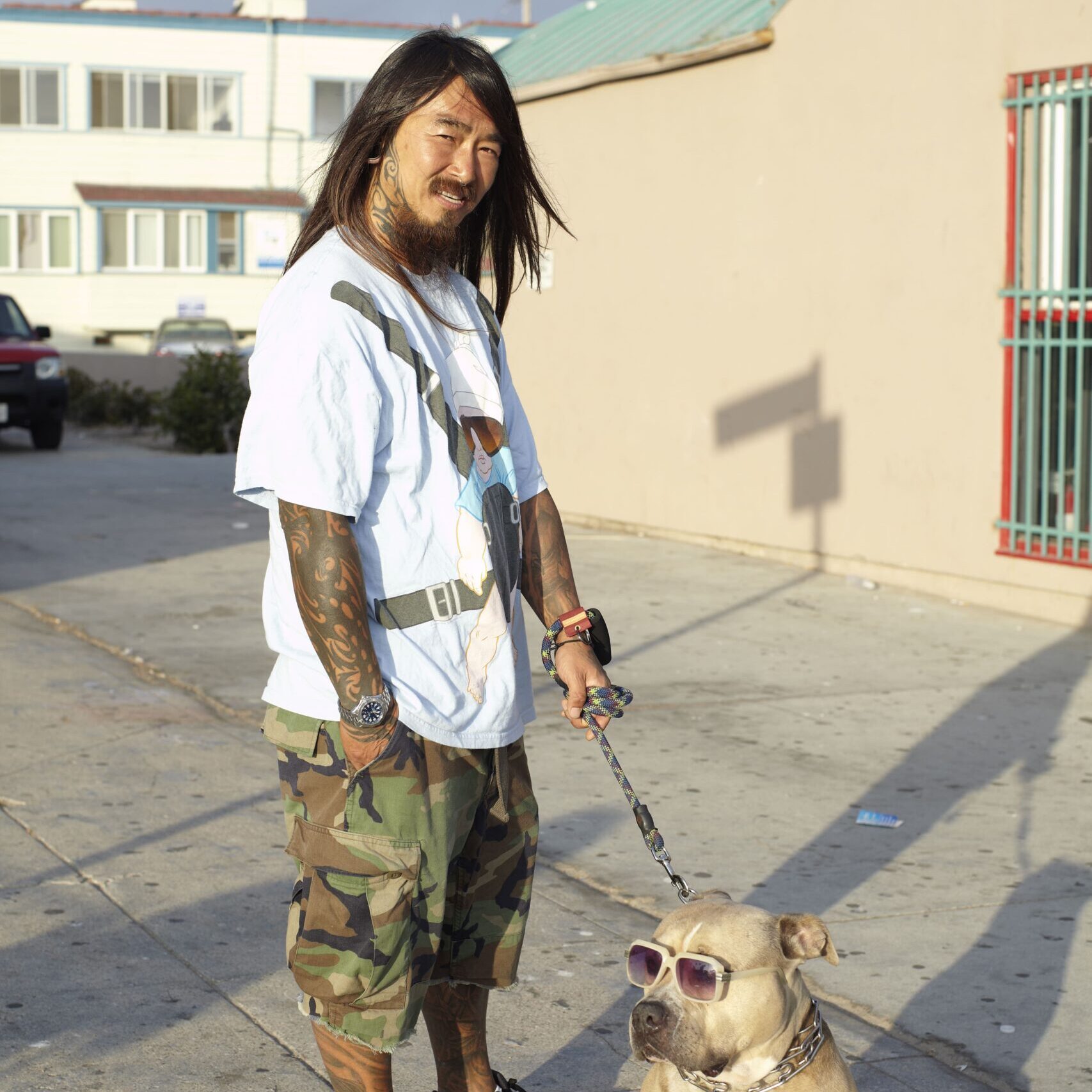 Man with long hair and tattoos stands on a sunny sidewalk, wearing a casual white t-shirt and camo shorts. He holds a leashed dog in sunglasses. Urban setting.