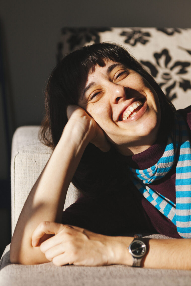 A woman with closed eyes and a joyful smile leans on a couch, wearing a striped scarf and a watch. Sunlight warmly illuminates her face, creating a relaxed atmosphere.