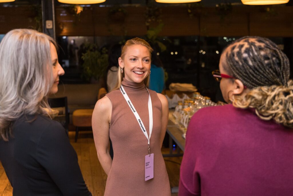 Three women conversing at an event, one in focus wearing a brown dress and name badge, smiling warmly. Cozy indoor setting with soft lighting.