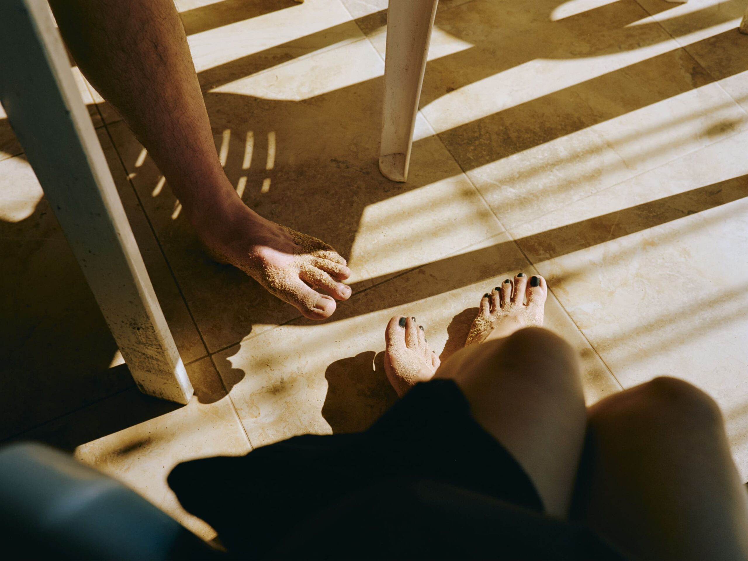 Two pairs of sandy bare feet rest on a sunlit tile floor, alongside chair legs casting striped shadows. Relaxed, warm ambiance.