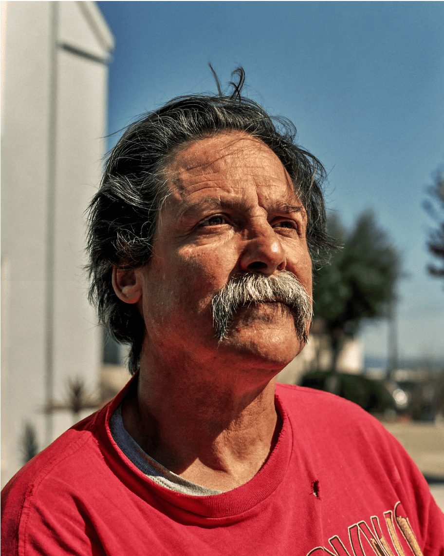 A man with a mustache and gray hair gazes upward, wearing a red shirt. The bright sunlight casts shadows on his face. A blurred outdoor background suggests a warm day.